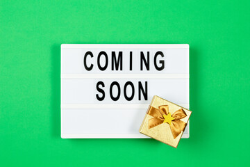 White lightbox with text COMING SOON and small gold present box on green color background. Top view