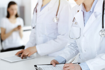 Unknown woman-doctors at work with patient at the background. Female physicians filling up medical documents or prescription while standing in sunny hospital reception desk, close-up. Health care