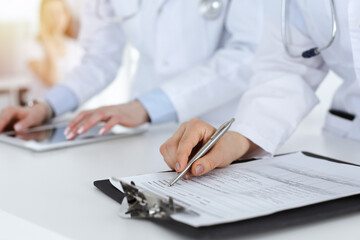 Unknown woman-doctors at work with patient at the background. Female physicians filling up medical documents or prescription while standing in hospital reception desk, close-up. Health care concept