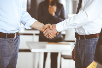 Fototapeta premium Two businessmen are shaking hands in office, close-up. Happy and excited business woman stands with raising hands at the background. Business people concept