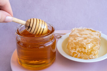 a woman's hand takes honey on a wooden stick from a full jar of honey with honeycombs on a plate