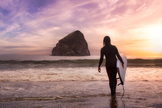 Adventurous Girl with a Surf Board is going surfing in the Ocean. Colorful Sunset Sky. Taken in Pacific City, Oregon Coast, United States of America.
