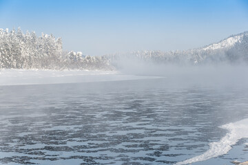 Frosty fog over winter river with snow and forest on bank. First ice on lake on cold day.