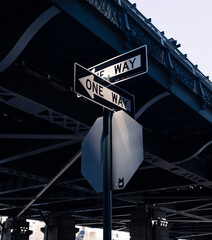 Two 'One Way' signs intersecting under a bridge in dramatic light