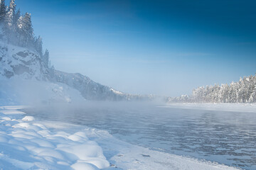 Frosty fog over winter river with snow and forest on bank. First ice on lake on cold day.