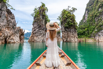 Back View of Young Female Tourist in Dress and Hat at Longtail Boat Exploring Lake with Limestone Cliffs at Cheow Lan Lake. Woman on Boat in Ratchaprapha Dam Khao Sok National Park in Thailand