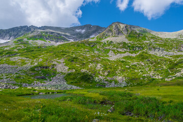 Rock ridge with snow and stone placers under blue sky. Summer trip to mountain valley. Atmospheric alpine landscape with stony meadow with green grass.