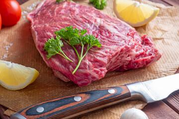 A piece of fresh raw beef lies on parchment surrounded by spices, herbs and vegetables. The knife is lying nearby. Side view. Food composition