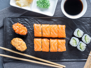 a variety of rolls and sushi gunkan nested on a black plate. next to it are bamboo wasabi sticks and sauce. Top view, flat lay. traditional Japanese cuisine