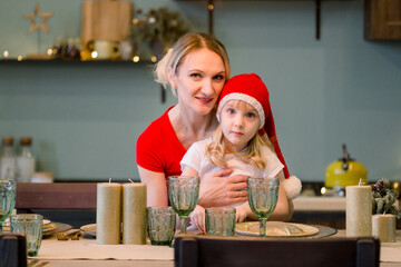 Mother with Happy cheerful Little girl in santa hat sitting at the festive table. Christmas time. New year 2021. Woman having fun with daughter