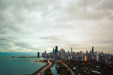 Naklejka premium Beautiful aerial skyline view of the city of Chicago with lights on the highrise and skyscraper buildings and traffic moving along Lake Shore Drive by the lake, zoo, and South Lagoon below at sunset.