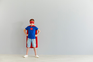 Serious little boy in superhero costume with mask standing at home over grey wall background.