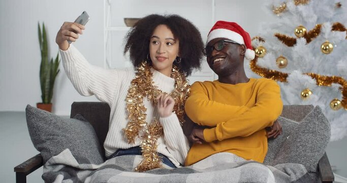 Young mixed race couple sitting on sofa in living room at home celebrating new year taking making holiday photo near Christmas tree filming video online for blog chatting in net video chat remotely