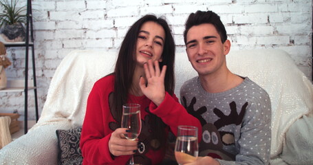 Attractive girl with boyfriend waving at camera sitting on sofa with glasses in hands