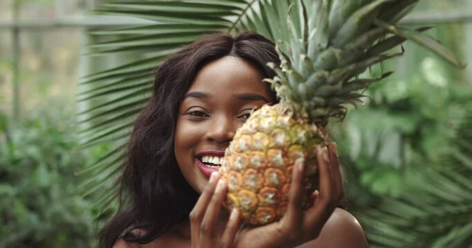 Young beautiful girl with natural makeup holds a pineapple in her hands and smiles at the camera. Against the background of tropical forest and wildlife. Shooting close up of emotion smile. The