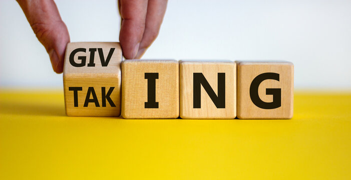 Taking or giving symbol. Hand turns a cube and changes the word 'taking' to 'giving' on wooden cubes. Beautiful yellow table, white background, copy space. Business and taking or giving concept.