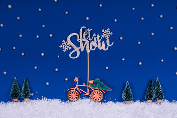 Funny cozy scene with Christmas toys on classic blue. Christmas tree is moved home by bicycle on the snow road with stars shining and Let Snow wording. Horizontal