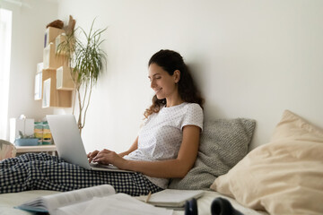 Smiling woman sitting on cozy bed with laptop on laps, enjoying leisure time, relaxing with gadget, shopping or chatting, satisfied happy young female working or studying online in bedroom
