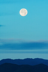 Full moon over some clouds and a celestial sky, and with mountains in the same color tone.