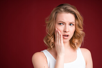 Blond woman with curly hair looks puzzled or having toothache in white tank top on red background.
