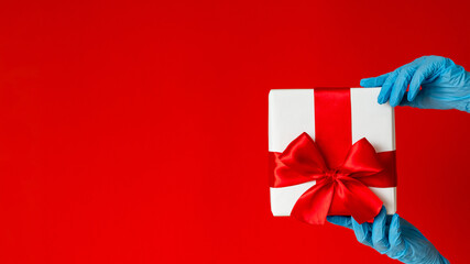 Gift surprise. Covid-19 restriction. Happy Valentine day. Greeting love. Hand in protective gloves holding present box wrapped taped ruby ribbon isolated on red copy space.