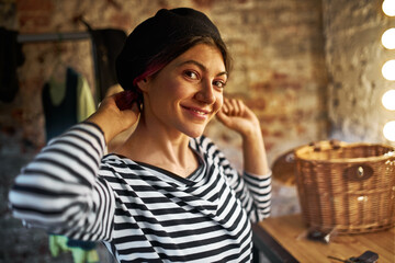 Half profile view of cheerful attractive young woman sitting in dressing room wearing stylish striped black and white shirt, vintage beret and nose piercing, looking at camera with happy smile