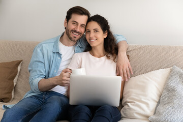 Portrait happy young couple sitting on couch with laptop, looking at camera, enjoying leisure time with gadget, shopping online, smiling husband holding cup of tea or coffee, hugging wife