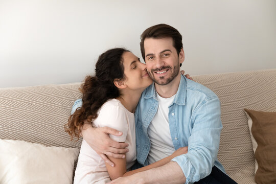 Close Up Loving Wife Kissing Husband On Cheek And Hugging, Young Family Enjoying Tender Moment, Happy Man Looking At Camera, Embracing Attractive Woman Girlfriend, Sitting On Cozy Couch At Home