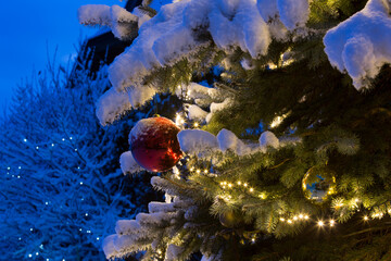 Snow covered christmas tree detail outdoor, with lights and red glossy ball x-mas ornaments.