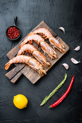 Fine selection of tiger prawns on cutting board with spices, pepper and lemon on black textured background, top view