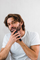 A man shaves his beard in the bathroom with an electric razor in front of a mirror. A handsome...