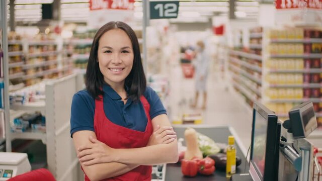 Medium close-up portrait of young smiling female cashier wearing red apron standing with hands folded in big mall while people doing shopping in background
