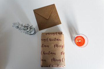 Close-up photo of a gift between a small Christmas tree and a candle with an envelope on top of a white table. During winter, thousands of gifts and Christmas cards are sent by post around the world.