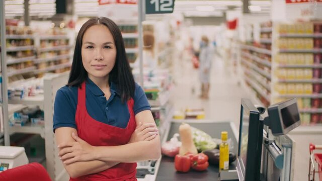 Medium close-up portrait of beautiful mixed-race woman working as cashier in hypermarket standing with hands folded with conveyor belt full of products in background