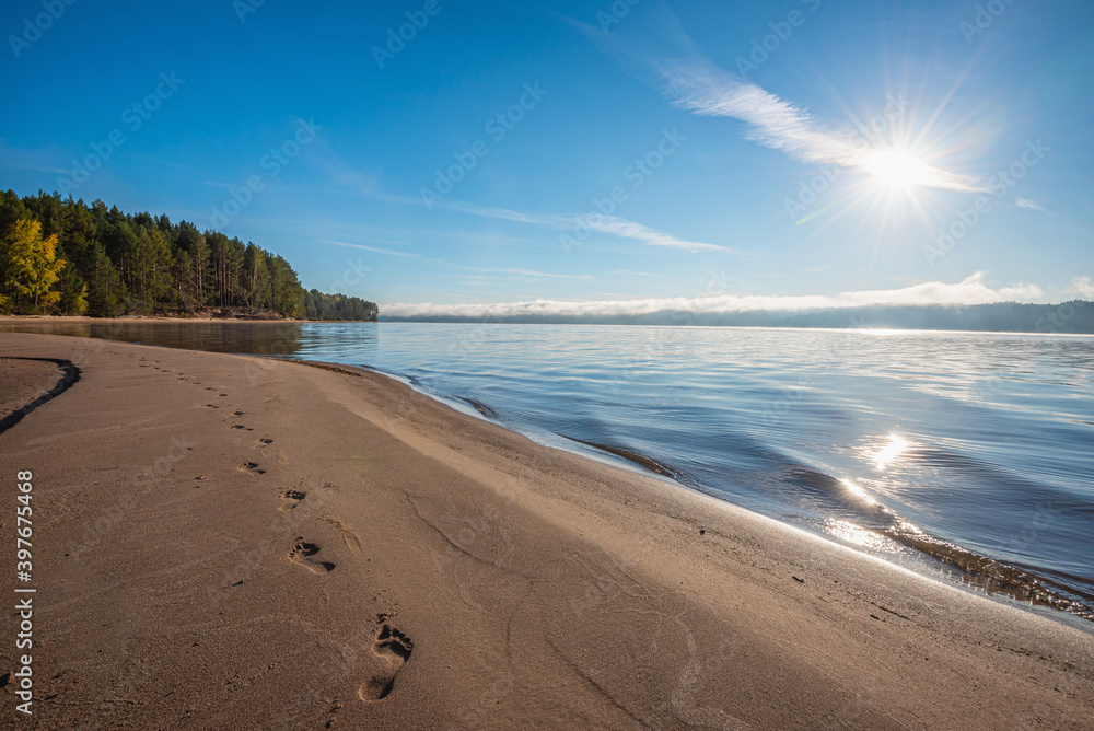 Wall mural bare footprints on the sand by the river, sunny morning and fog over the water, sun rays.