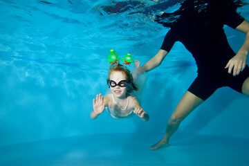A little girl in a dress is swimming under the water in the pool. Christmas decorations on the...