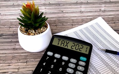 Taxes in 2021 on the calculator.Next to it is a pot with a flower, a pen, and a document with numbers on a wooden background .