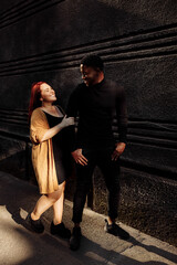 Fashion couple walking at the city, stylish woman cuddle with attractive African American man, adorable wife spend weekends with caring husband, street photoshoot concept