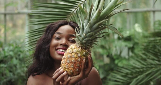 Young beautiful girl with natural makeup holds a pineapple in her hands and smiles at the camera. Against the background of tropical forest and wildlife. The girl covers her face with a pineapple. The