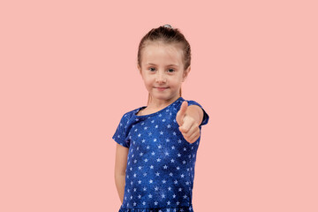 A beautiful young brunette girl in a blue dress shows a thumbs up gesture. Pink isolated background.
