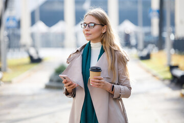 Cheerful young woman wearing coat walking with coffee cup and cellphone