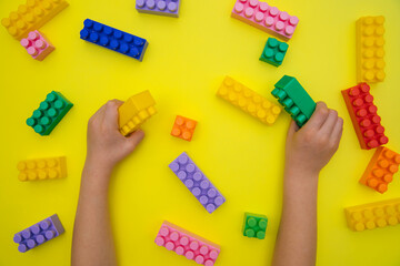 Children's hands hold the details of the constructor on a yellow background. Multi-colored construction blocks.