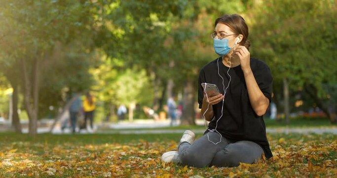 Beautiful girl sits on lawn in autumn city park and looks photos in his smartphone. Then she takes off and throws out mask and continues to looks photos without it. Cinema 4K 60fps video