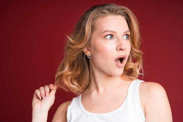 Blond woman with curly hair in white tank top looks shocked on red background.