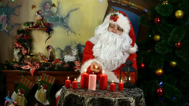 Jolly Santa set fire candles while sitting by a Christmas tree decorated with Christmas balls and beautiful decorations and rings a bell and dances. Medium plan.