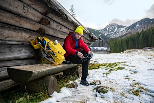 Hiker man in a yellow cap and a red down jacket sits on a wooden bench and puts crampons on his boots. The yellow backpack is lying next to the tourist.