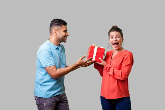 Best present for woman. Young man in casual clothes giving gift box to pleasantly surprised woman looking at camera with open mouth, unexpected bonus. isolated on gray background, indoor studio shot