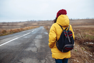 Obraz na płótnie Canvas Backview of a female tourist with a backpack wearing yellow jacket and red hat walks on the road. Young woman travels during winter or late autumn season. Hitch-hiking, trip and travelling concept.