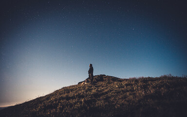 Fototapeta premium Hiker standing on top of a mountain and enjoying night sky view with a lots of stars and artificial city lights