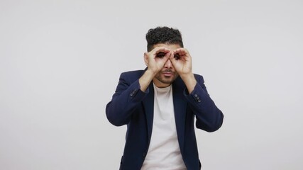 Funny bearded man in suit spying holding fists near eyes and looking through holes, pretending to watch in binoculars, zooming to see better, having fun. Indoor studio shot isolated on gray background - Powered by Adobe